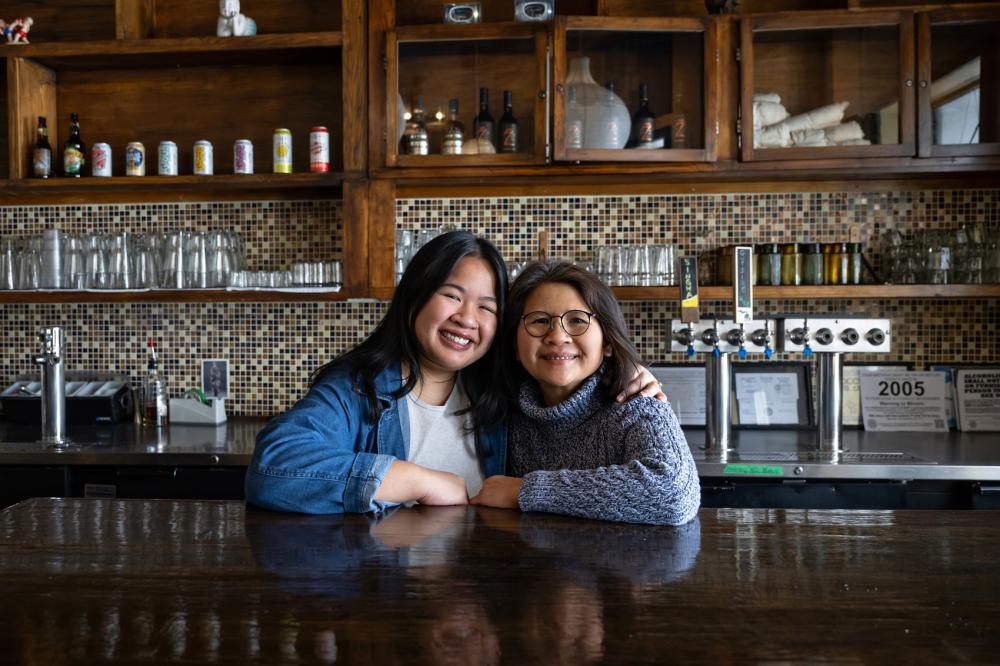 An adult daughter and mother smile for the camera, standing behind a counter inside of a restaurant