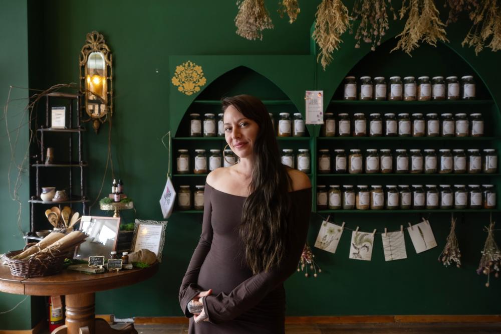 Person stands inside of a store and smiles and poses with their arms posed to the side, wearing a brown, strapless long sleeve dress