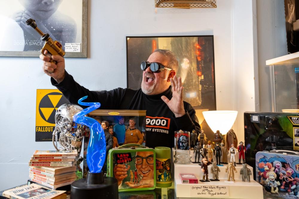 Man stands behind counter in an antique and retro shop. He's wearing goggles and holding a toy laser blaster. In front of him on the counter are toys and memorabilia from tv shows.