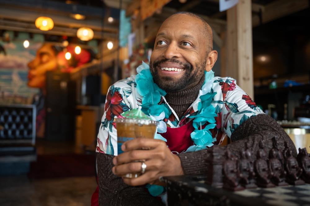 Man holds coffee in plastic cup as he smiles and looks to the left. The man is wearing a blue andd red hawaiian shirt and sitting at a chess table inside a cafe.