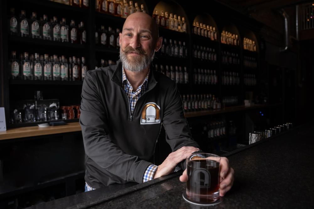 Man smiles and stands behind a bar counter as he holds a glass full of a beverage, almost as if he is inviting you to take the glass from him.
