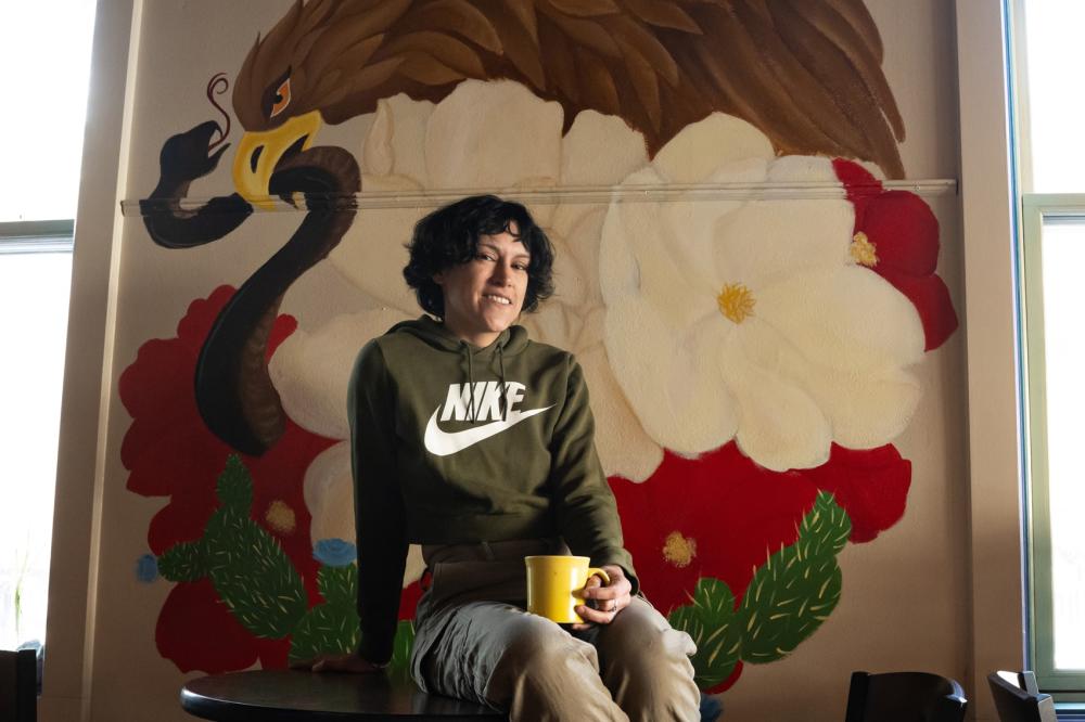 Woman poses and smiles for the camera while sitting on table and holding a yellow coffee mug. A large mural of an eagle catching a snake, surrounded by flowers is painted on the wall behind her.
