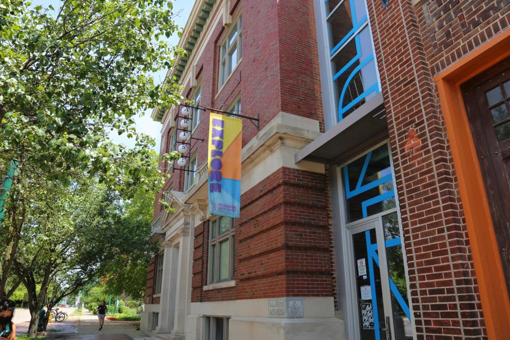 Riverside Arts Center exterior building with 'explore' banner on building flag. Green trees line the…