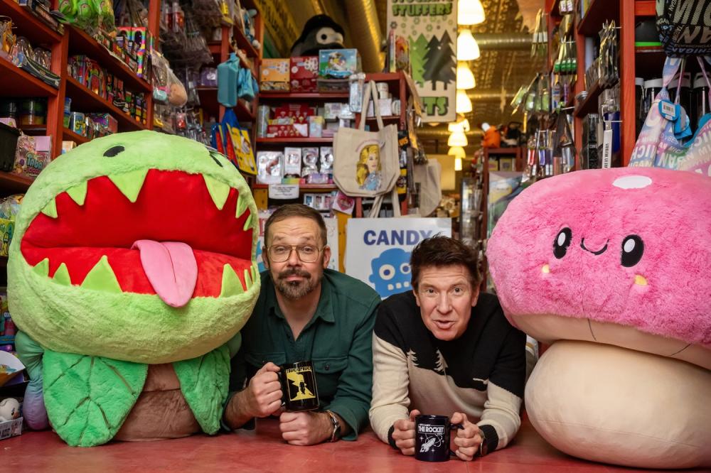Two men pose between two large giant stuffed animals. The men are each holding a novelty mug in their hands. Behind them are aisles of candy and novelty gifts.