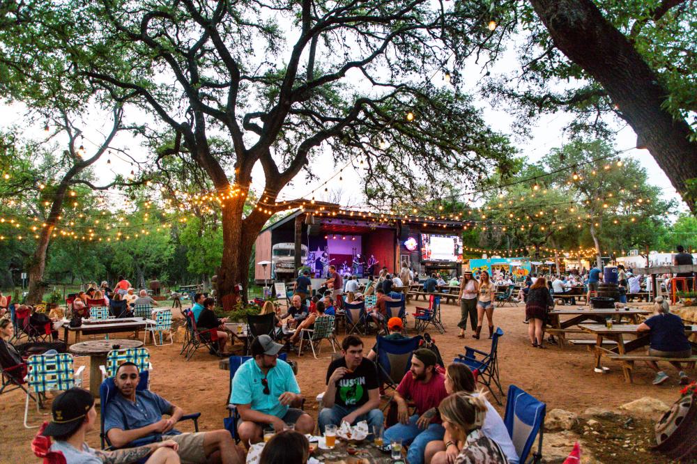 People sitting in law chairs chatting with one another at Armadillo Den with string lights hung from the trees and a stage behind them.