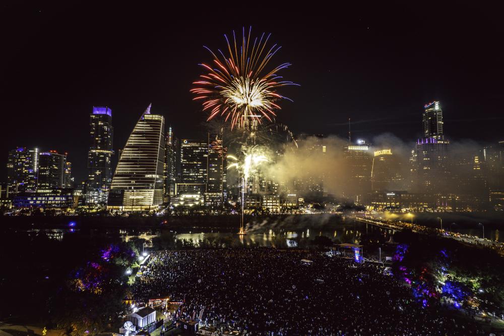 Red fireworks explodes over downtown Austin skyline and Lady Bird Lake at night. Aerial view shows a music stage and crowd gathered below at Auditorium shores.