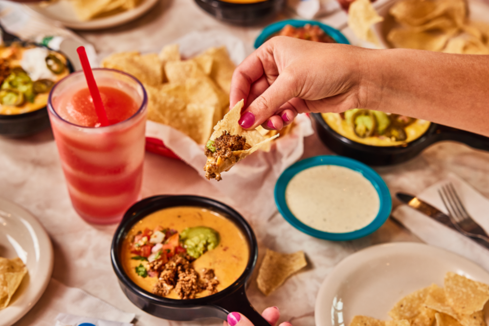 A table at Chuy's filled with queso and chips with someone's hand reaching down into the frame to scoop queso with a chip.