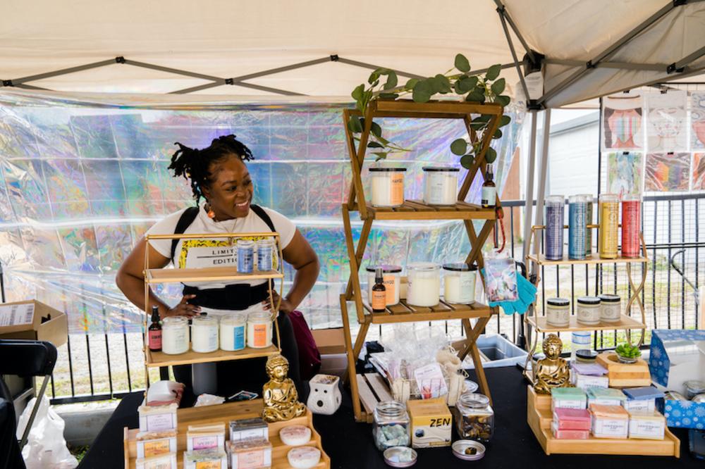 Woman standing behind vendor booth with homemade candles and soaps.