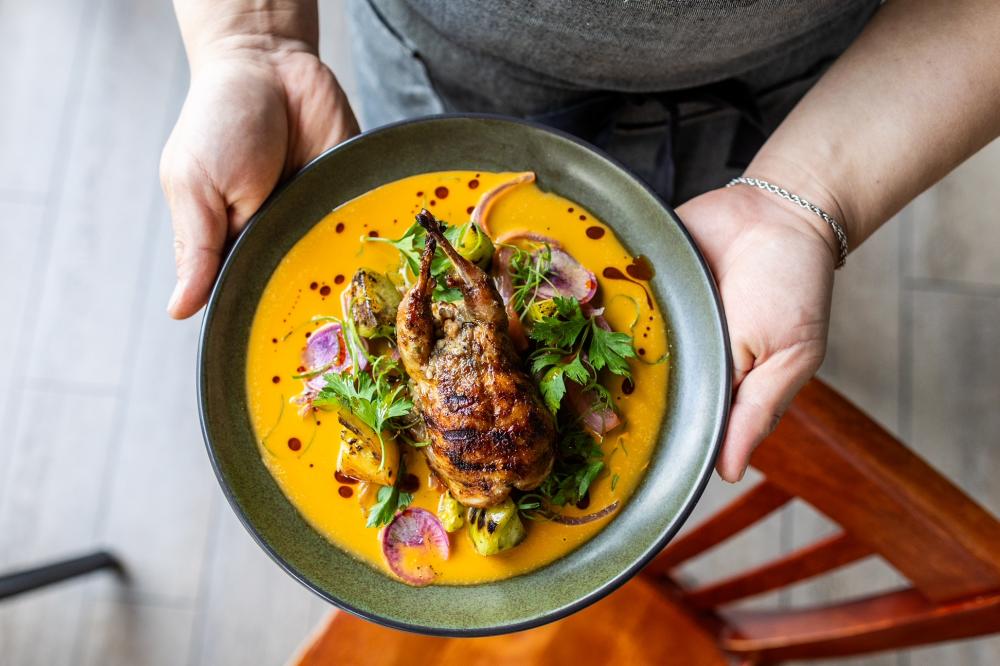 Hands holding a plate covered in a yellow-orange sauce, colorful greens, and a perfectly roasted quail.
