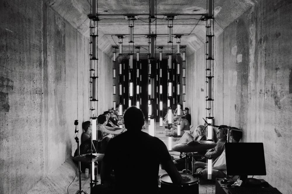 Black and white image of concrete room lined with mirrors and lights, with an audience on the floor and drummer playing a set.