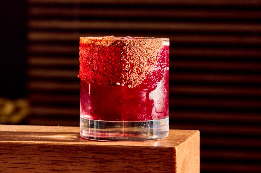 A red margarita with tajin on the rim of the glass sitting on the edge of a wooden table.