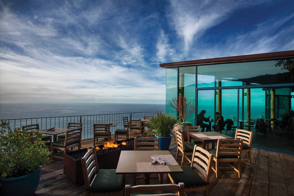 This is an image of Sierra Mar Restaurant at Post Ranch Inn in Big Sur taken from the patio. A fireplace is lit on the patio, surrounded by wooden tables and chair. The vast blue ocean can be seen in the background as well as the glass structure of the restaurant