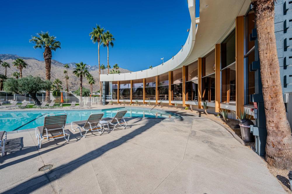 A modern resort swimming pool with lounge chairs, surrounded by tall palm trees, with a mountain backdrop under a clear blue sky.