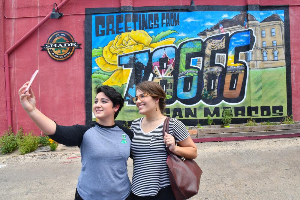Couple takes a selfie in front of the Greetings from 78666 mural in Kissing Alley in downtown San Marcos.