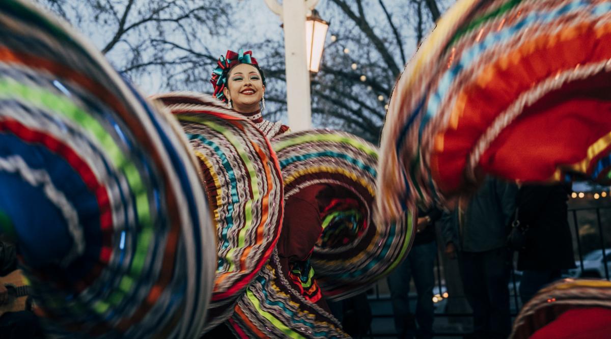 A performer in vibrant traditional Mexican attire, including a colorful headscarf, smiles while twirling the large, multicolored skirt during a cultural dance performance.