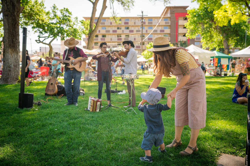 A woman holds the hands of a young child as they dance to a live band at the Downtown Growers Market
