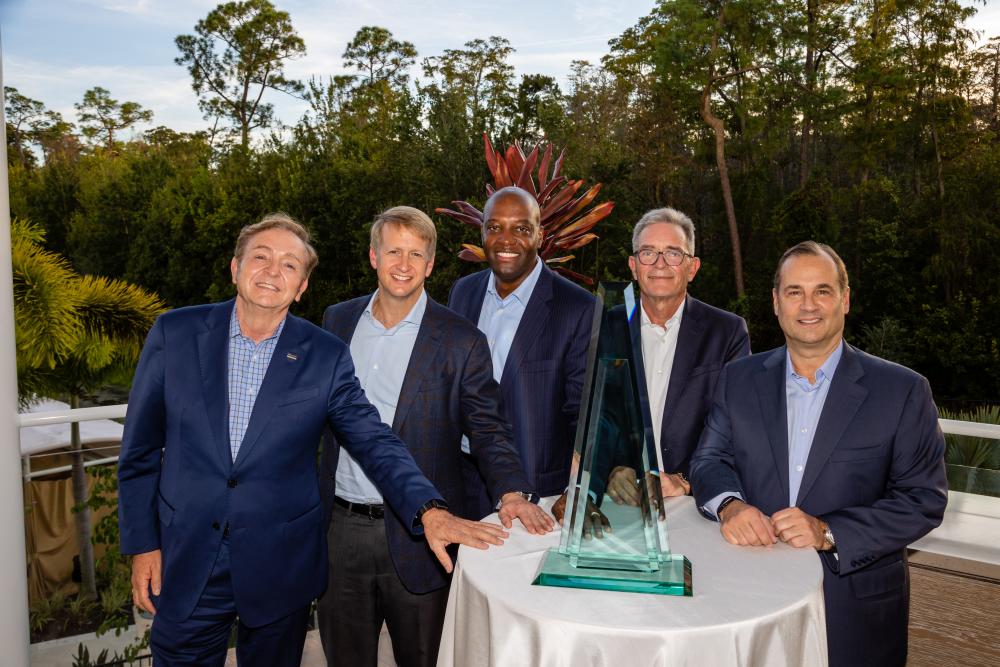 Image of five gentlemen in suits standing next a table draped with white linen. On top of the table is a glass triangular shaped award.