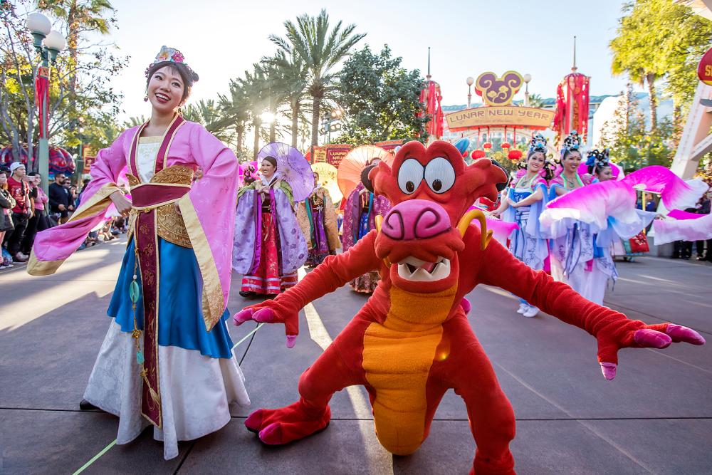 Image of a woman, Mulan, dressed in a pink, blue, and white gown dancing inside Disney California Adventure Park. Standing next to her is a human dressed as Mushu (a red dragon) from Disney's Mulan. They are apart of a parade taking place.
