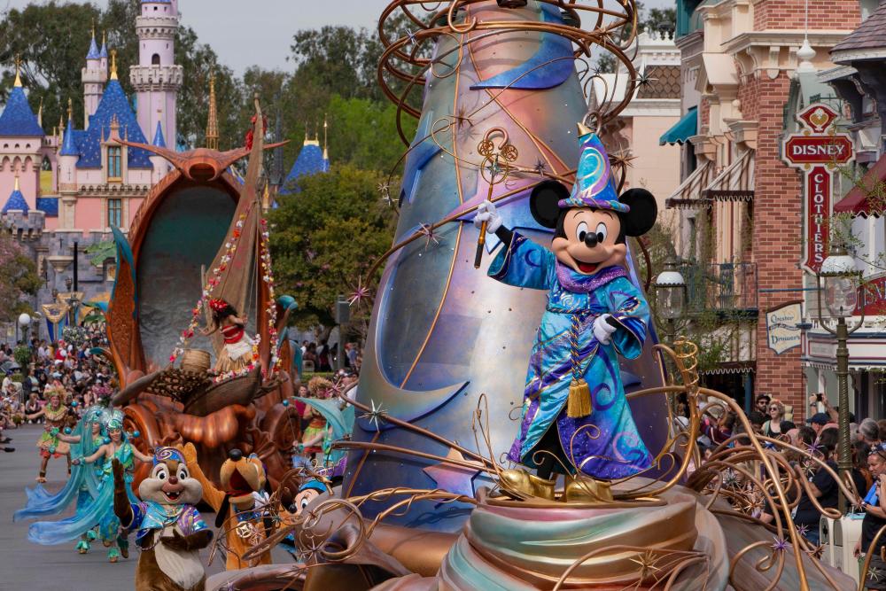 Image of a blue, purple, and pink structure floating down Main Street U.S.A. inside Disneyland. Mickey Mouse can be seen standing on the structure and wearing a blue and purple wizard outfit.