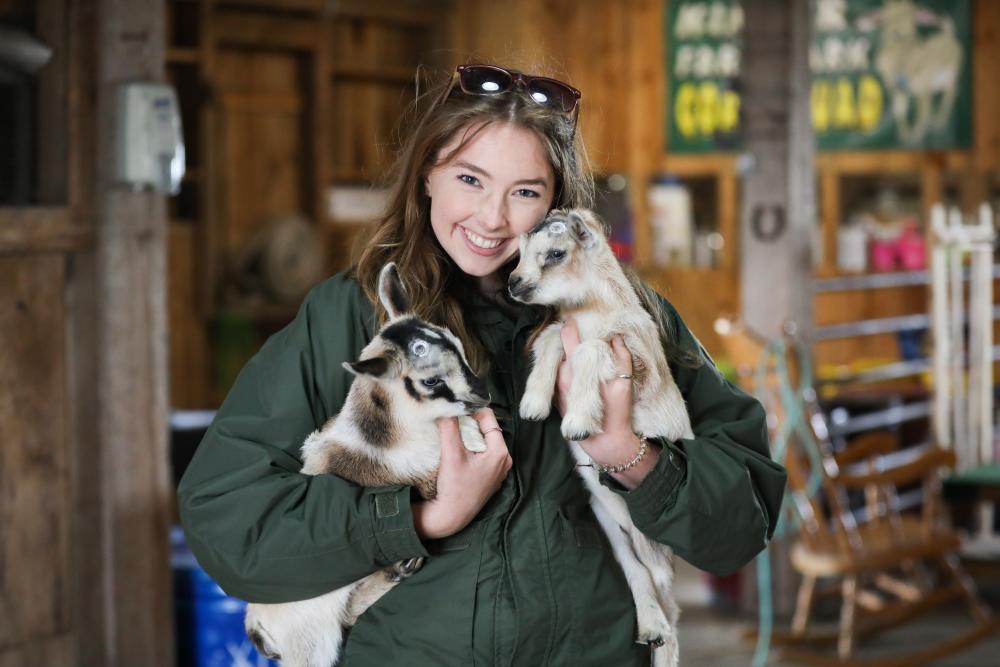 A woman smiles and holds up two baby goats in a barn