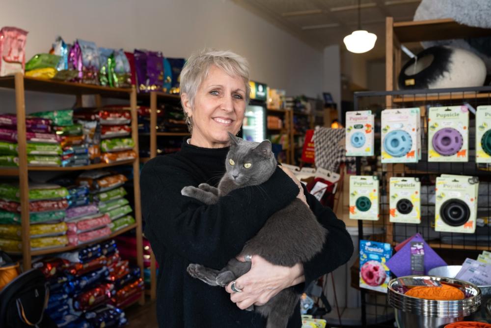 Woman wears a black sweater and is holding a grey cat in her arms. She smiles and looks at the camera. Behind her are shelves of pet food supplies and pet toys.