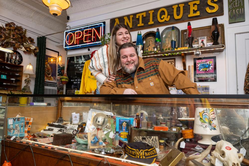 Woman leans on man and they both smile as they smile and post for the camera. They sit behind a counter that is full of antique and vintage finds. Behind them on the wall, there is a light up "open" sign, and another wooden sign that reads "antiques"
