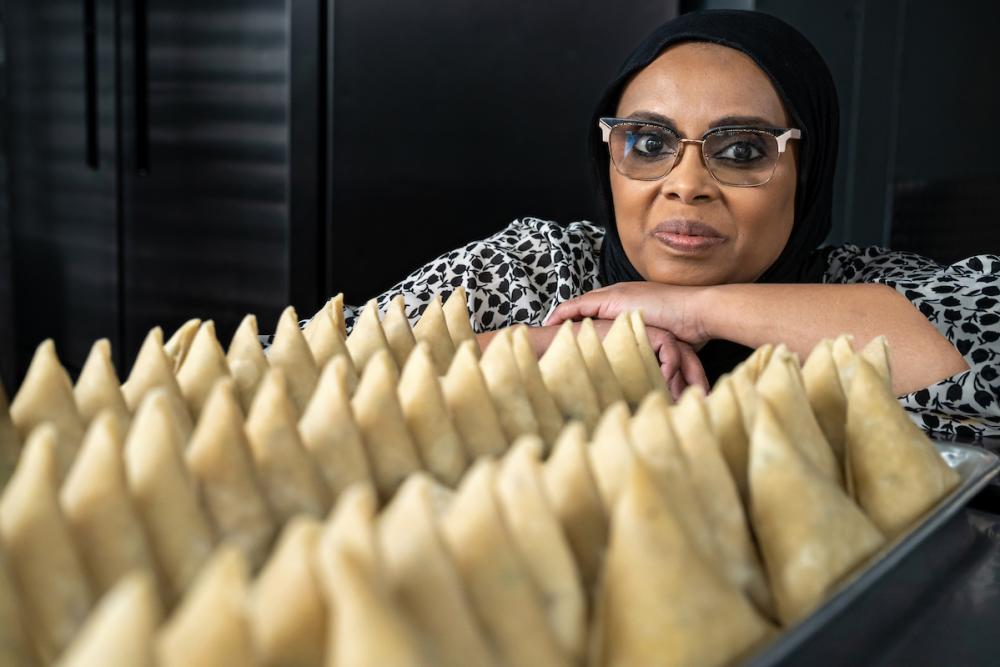 Woman smiles at the camera while resting her head on her crossed arms. In front of her are three rows of dozens of samosas.