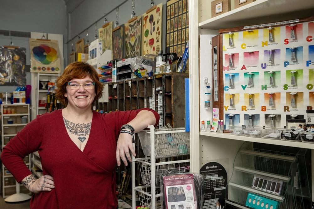 woman wearing a red shirt smiles and poses for a photo. She stands inside an art shop full of supplies for sale.