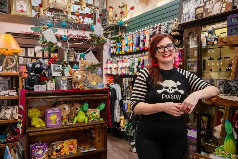 woman stands inside of retail shop. She wears a black and white shirt that reads "poison" below a skull and crossbones design. Behind her in the shop are individual items placed on shelves and hanging from fixtures.