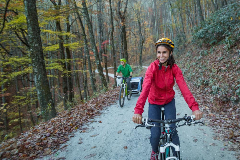 Woman wearing pink jacket riding bike on trail with husband behind during fall