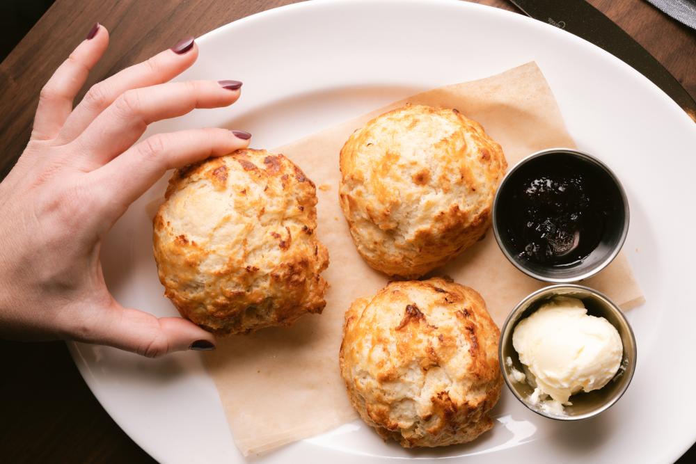 Photo of a hand holding a homemade biscuit from Corinne restaurant, there are two other biscuits on the plate along with a silver bowl of butter and another silver bowl with a dark jam