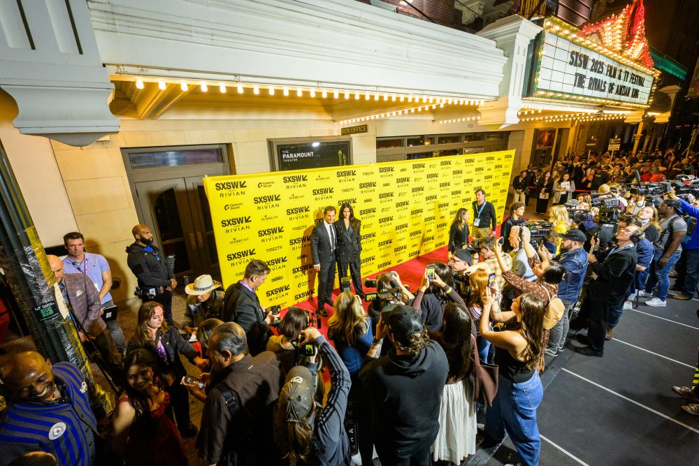 A photo looking down onto the red carpet out front of the Paramount Theatre during SXSW with two people posing on the carpet and photographers snapping photos.