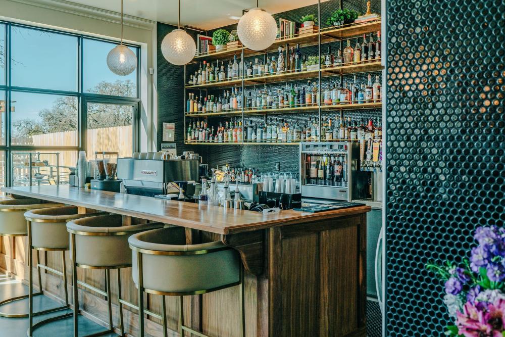 Speakeasy-style bar with decorative pendant lighting, liquor-stocked shelves, and teal-hexagon-tiled wall.