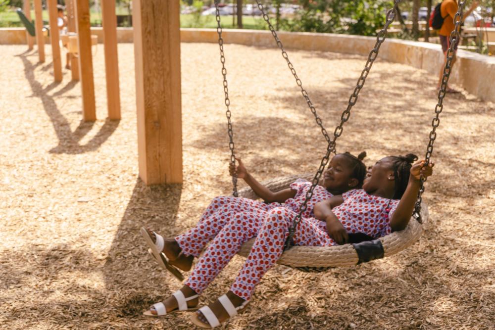 Two kids laughing on swing together in matching outfits at a local park.