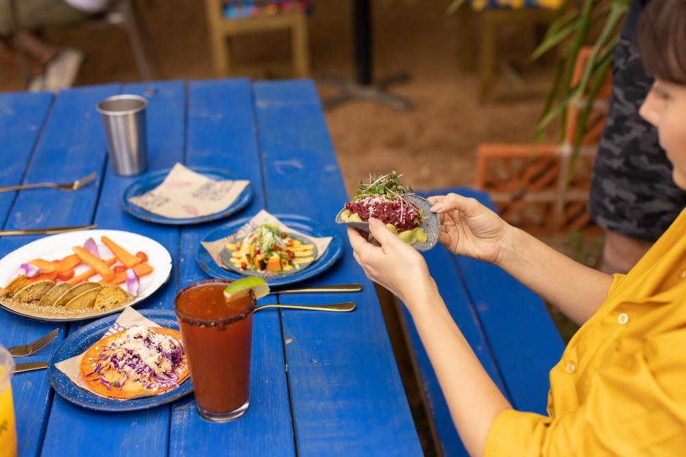 Woman sitting at bright blue picnic table, picking up a very full taco in a blue corn tortilla.