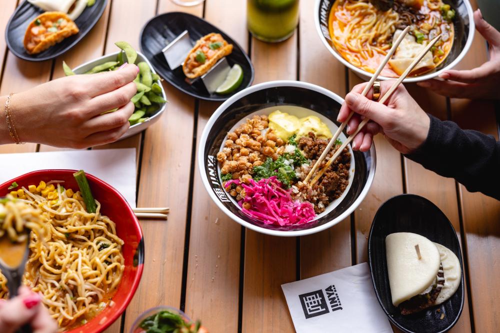 Spread of Asian-fusion foods on a wooden table with hands and chop sticks picking items off of plates and bowls.