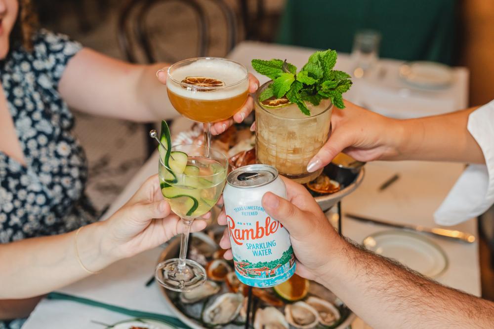 Photo shows four people's hands holding cocktails and Rambler sparkling water in a cheers position