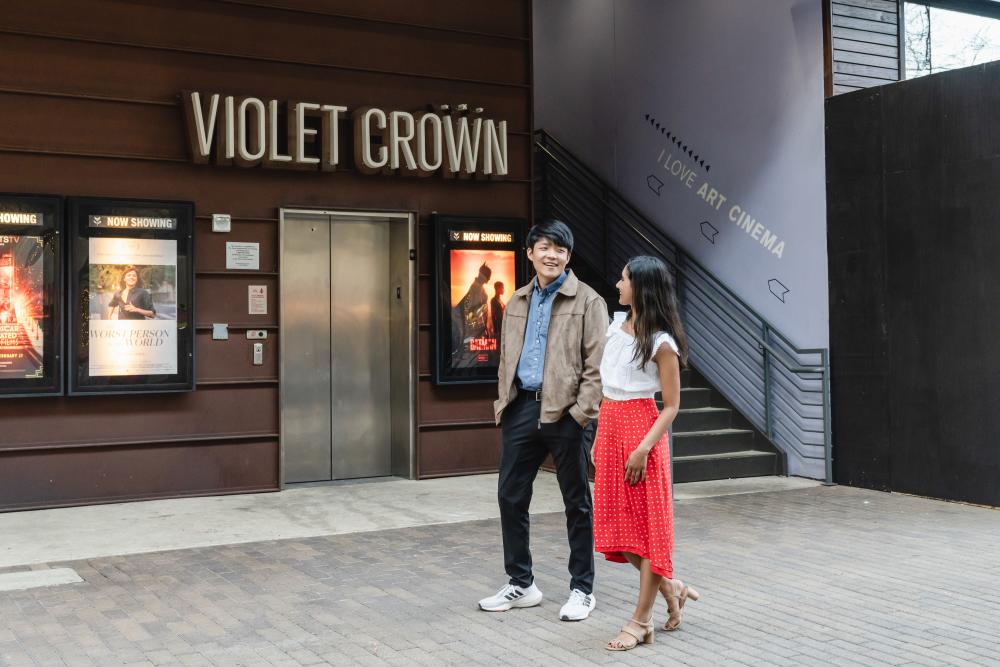 man and woman walking down sidewak in front of movie theater posters, elevator and sign reading Violet Crowd