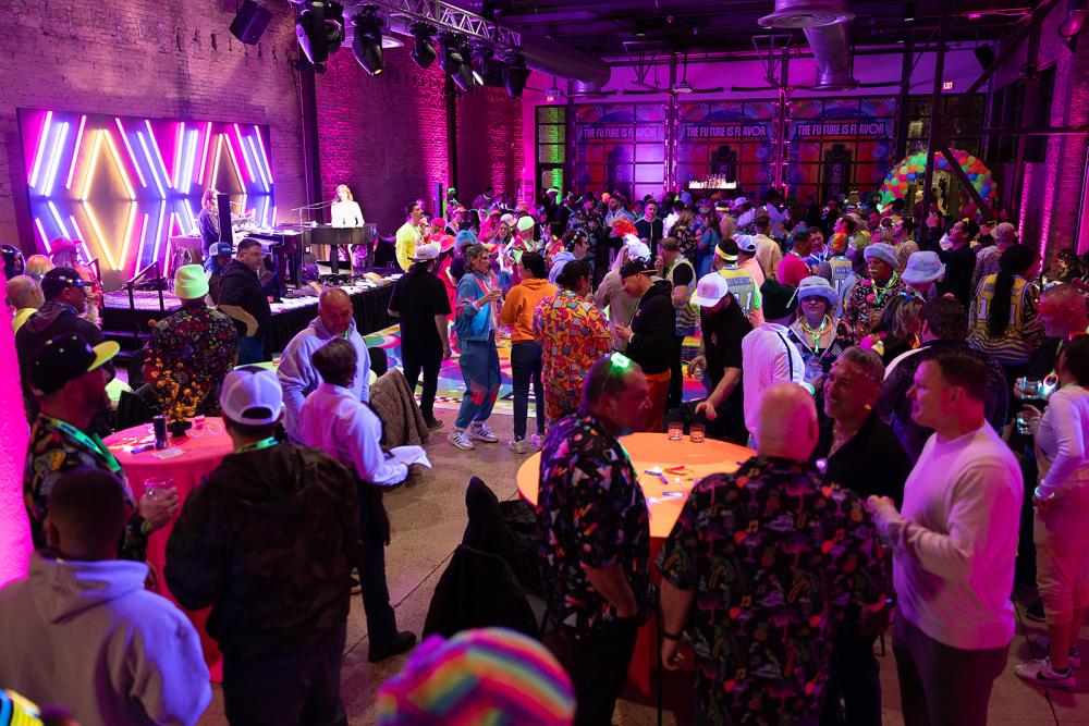 Group of people in a venue with a neon backdrop on a stage, enjoying a conference after party.
