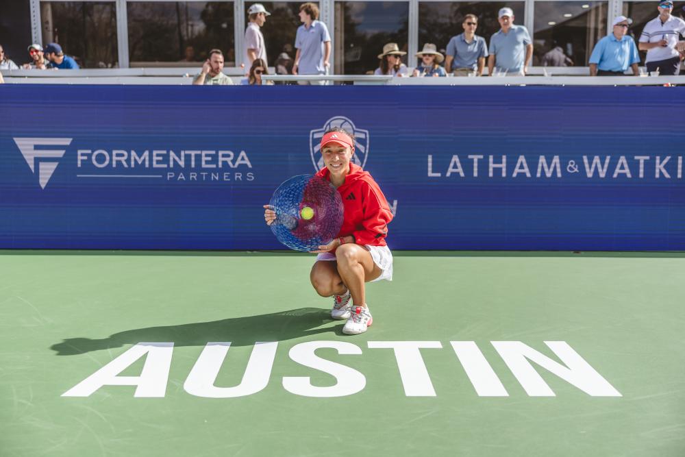 Jessica Pegula holding the ATX Open 2025 trophy and squatting on the ground in front of the bold, white "Austin" text printed on the green tennis court surface and smiling at the camera.