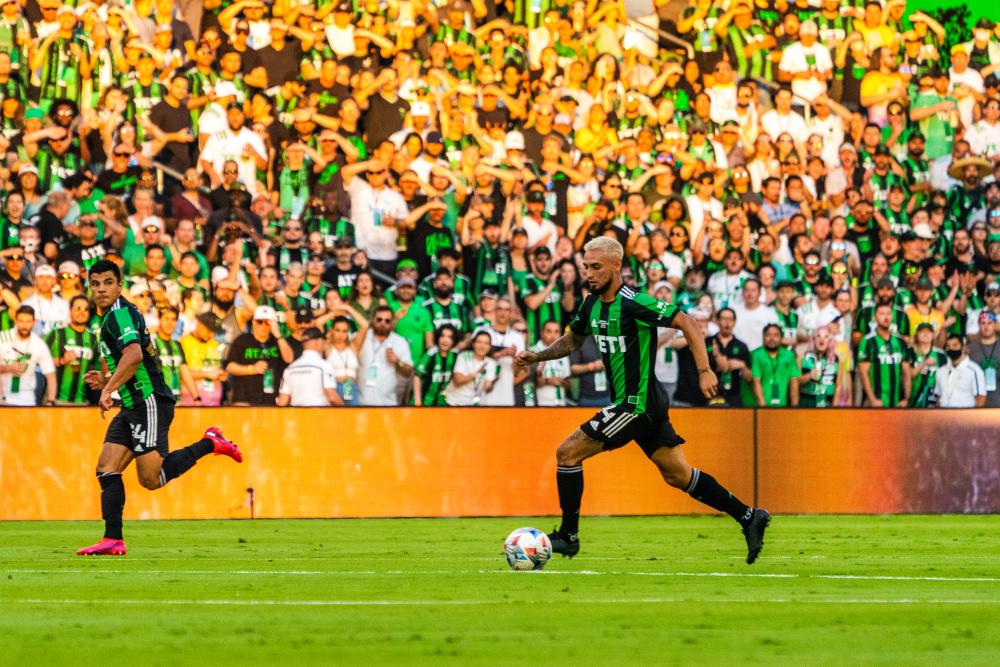Two Austin FC players running the same direction while the sun shines bright on a cheering crowd in verde shirts.