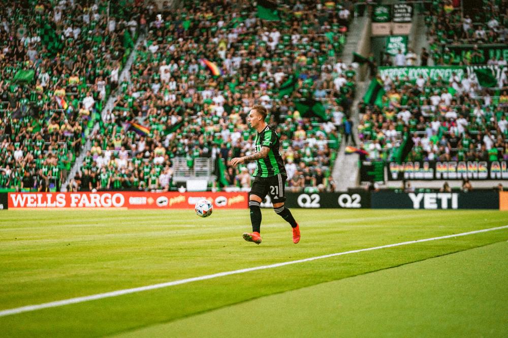 Žan Kolmanič for Austin FC on the field with the ball at Q2 stadium. A large crowd in the Supporters Section is visible behind him, waving rainbow flags