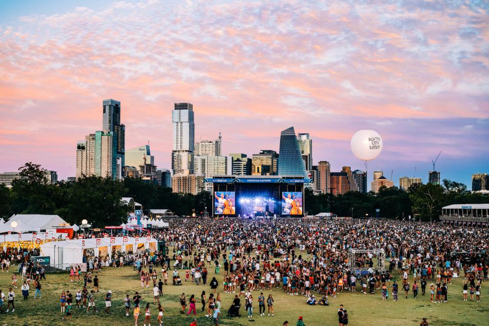 Aerial shot of Zilker Park during ACL festival with a sea of fans crowding the main stage, a cotton candy colored sky with the Austin skyline as the backdrop.