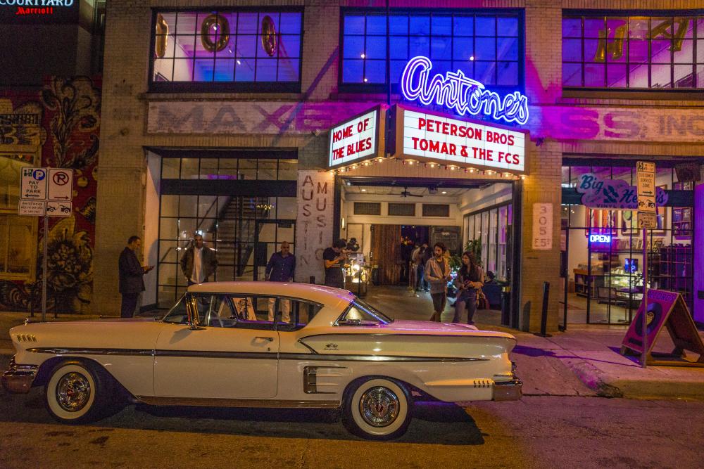 People buzzing in and out of Antone's at night. The iconic neon sign is lit and a vintage car is parked out front of the venue.