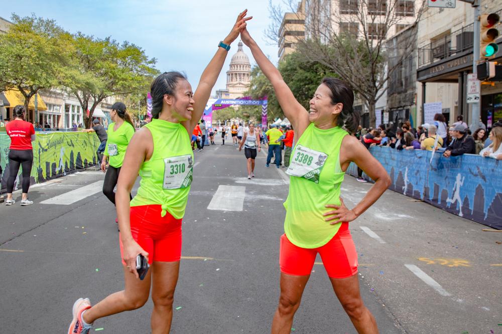 Two women in matching athletic clothes high-fiving in front of the Texas State Capitol Building and finish line of the Austin Marathon.