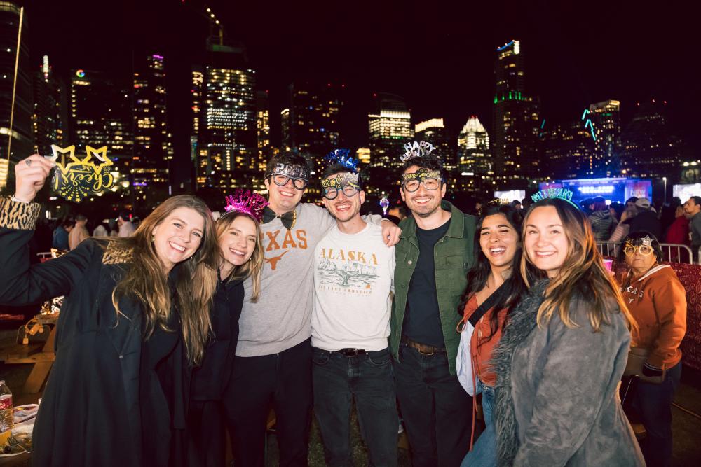 Group of friends posing for a picture in their New Year accessories in front of the Austin skyline at night.