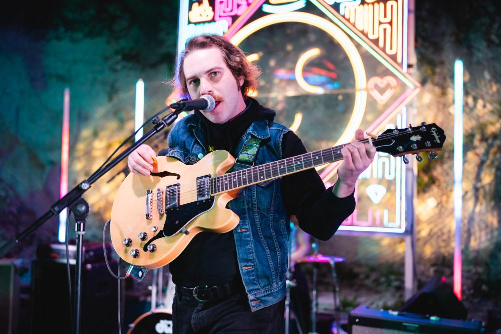 Man with electric guitar singing into a microphone with Cheer Up Charlies' neon stage background behind him.