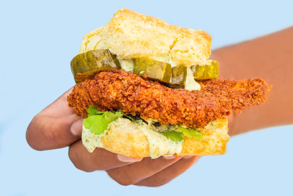 A hand holds a fried chicken biscuit sandwich with pickles, lettuce and dripping sauce at Bird Bird Biscuit