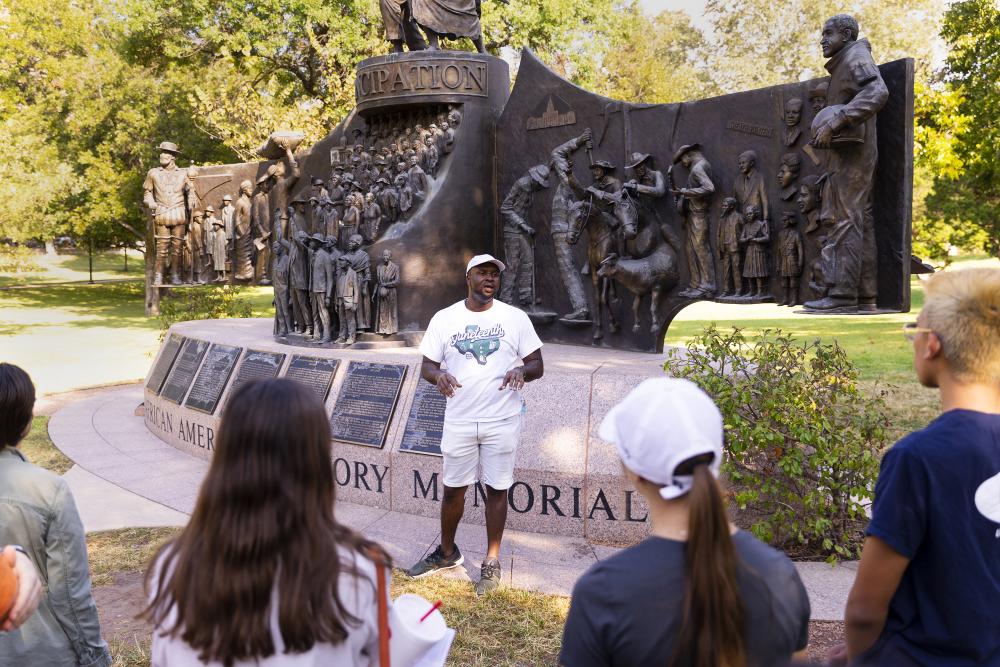 Javiar Wallace, founder of Black Austin Tours, standing in front of a memorial statue, talking to a group of people giving a tour.