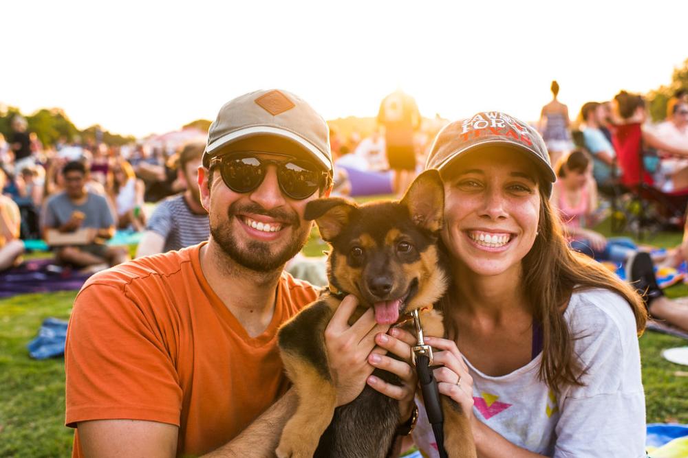 A man and woman holding a german shepherd puppy smile at the camera during ACL Radio's Blues on the Green. They are sitting on a blanket in Zilker Park